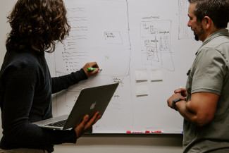 Two people writing on a board, one of them holding a laptop